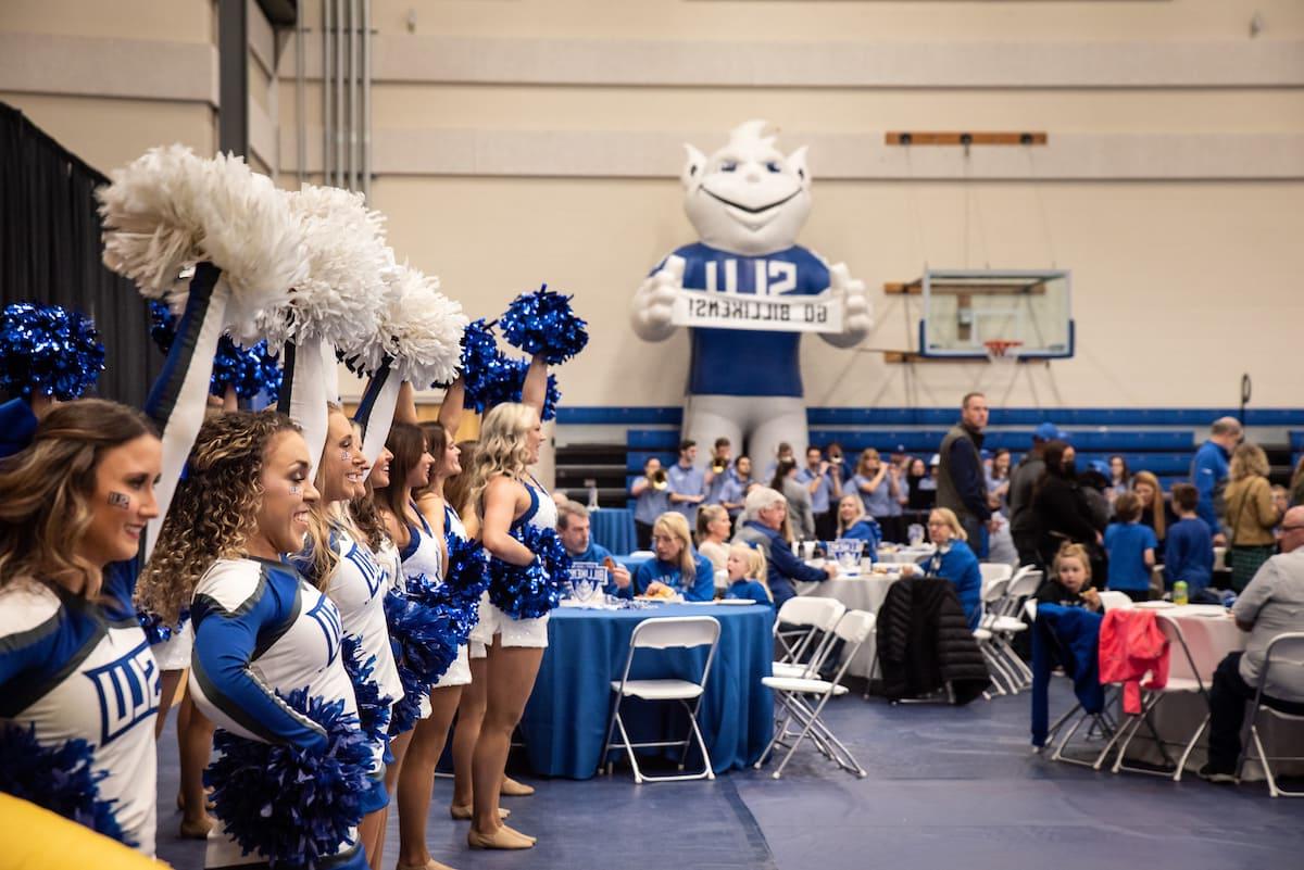 Members of the 博彩网址大全 cheer and dance team stand in a line and smile, holding blue and white pom poms in the air. 在左边, 真正的蓝粉盛会 visitors wearing blue and white 博彩网址大全 spiritwear sit around tables adorned with "Saint 路易 Billikens" signs. 在后台, members of the 博彩网址大全 pep band ready their instruments in front of a basketball hoop and a large inflatable Billiken holding a sign that reads "Go Billikens!"