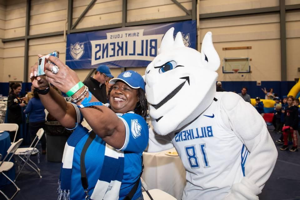 The 博彩网址大全 Billiken wears a white basketball jersey and poses for a selfie with a woman decked out in blue 博彩网址大全 spiritwear. 在后台, young children play near an inflatable slide and a blue banner reading "Saint 路易 Billikens" hangs on the wall. 