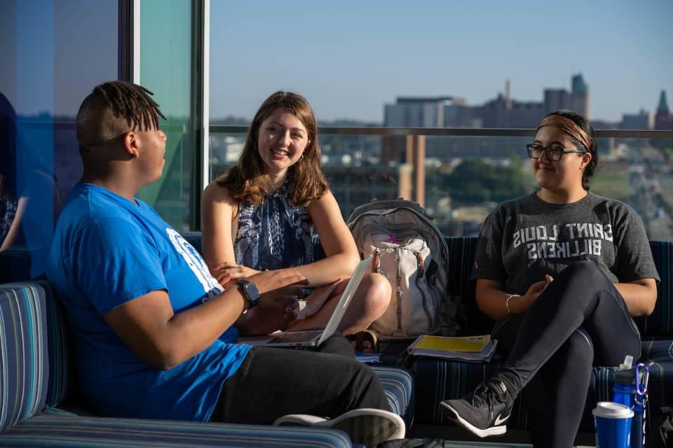 Three students chat in a sunny spot in a common area in Grand Hall.