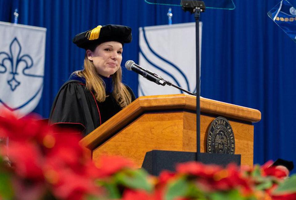 Dr. Katie Kelting offers remarks to the graduates from behind the commencment stage inside a crowded Chaifetz Arena.