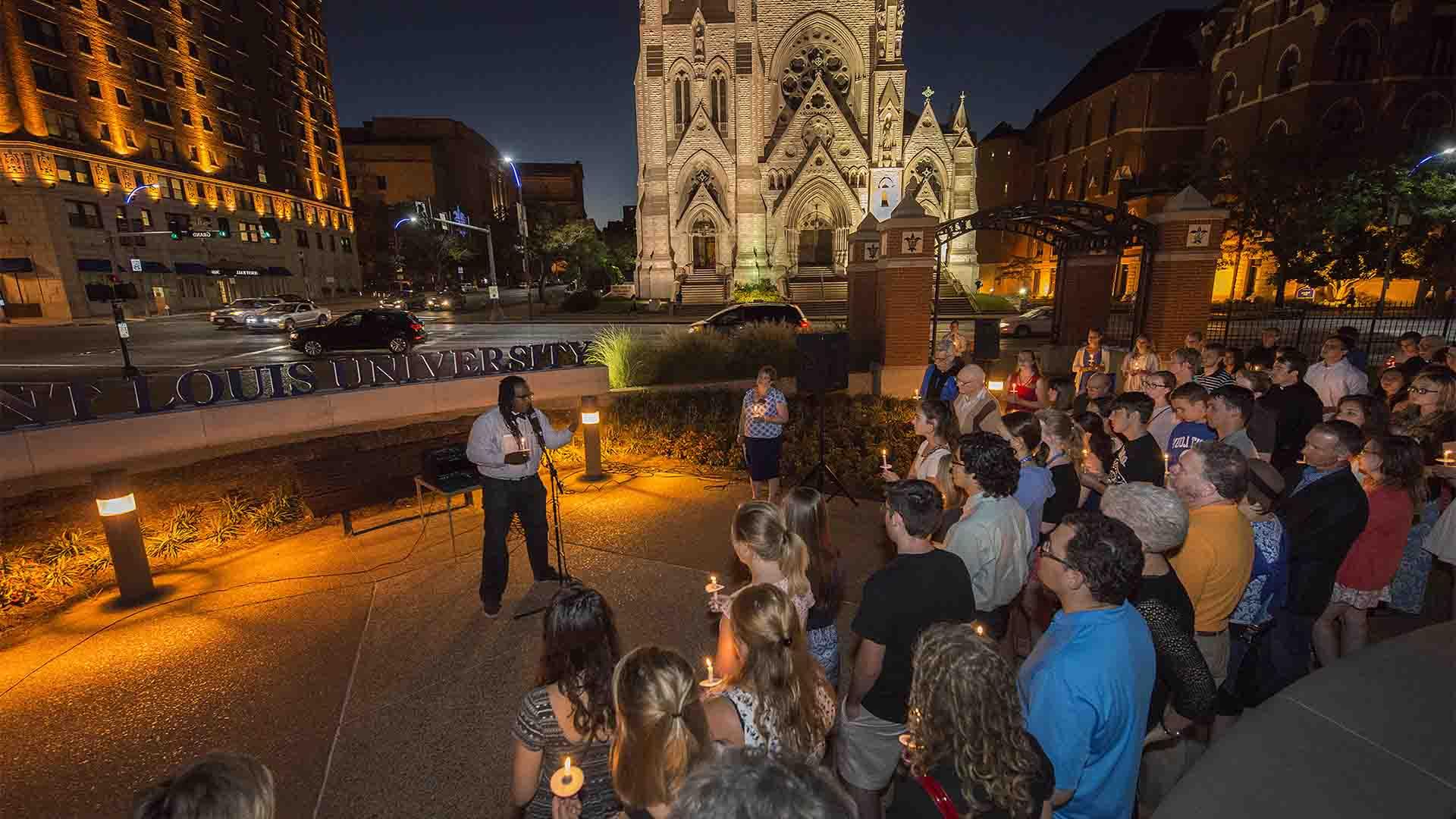 Dr. Jonathan Smith stands in front of a group of people at night, at the corner or Lindell and Grand, with the Saint Louis University sign in the background.