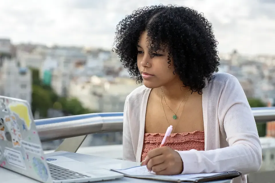 Student taking notes on a notepad and looking at a laptop while sitting at a table on an outdoor balcony.