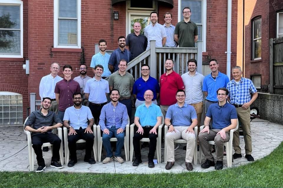 A group of residents pose for a photo in the back of the Jesuit house, some sitting on lawn chairs, and some standing in a row behind and on steps.